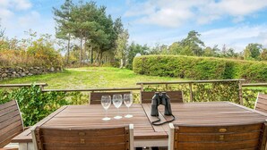 Een houten buitentafel met vier stoelen, drie wijnglazen en een verrekijker op een terras met uitzicht op een aangelegde tuin en bomen.