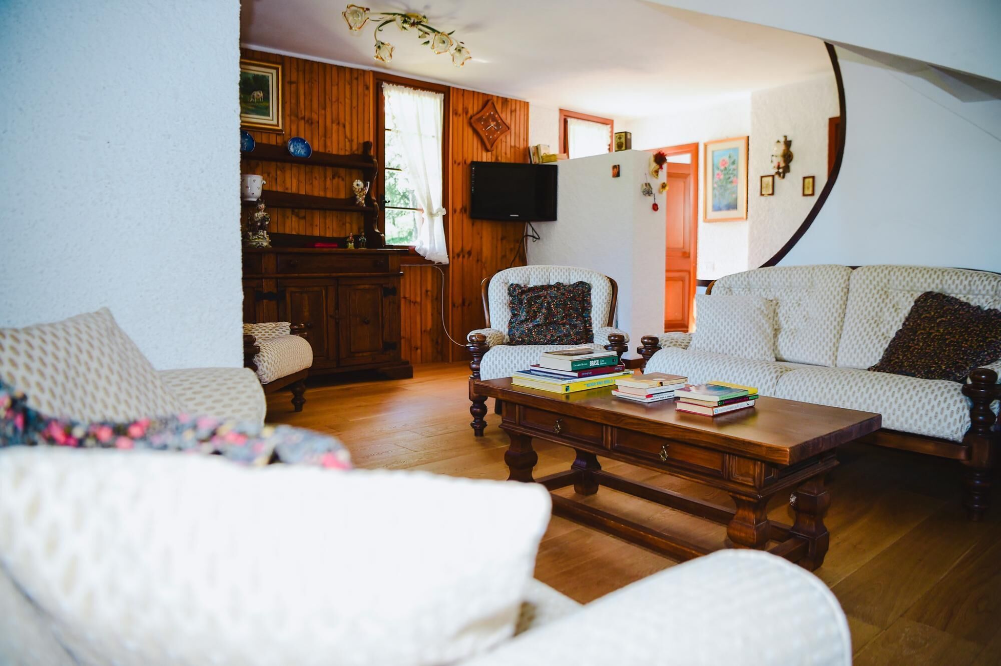 A cozy living room with a wooden floor, a white sofa, a patterned armchair, and a coffee table with books.