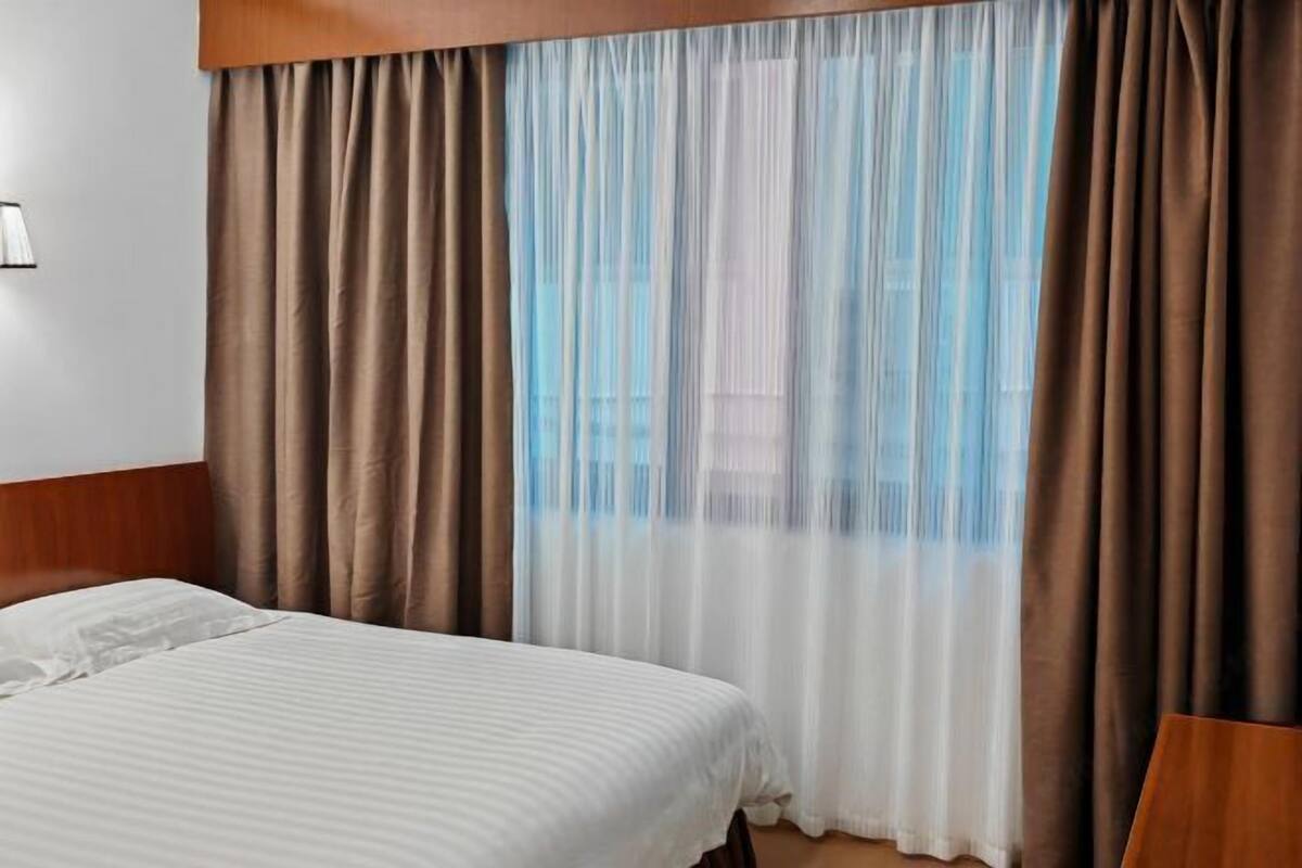 Habitación de hotel con una cama, una ventana con cortinas traslúcidas y cabecero de madera.