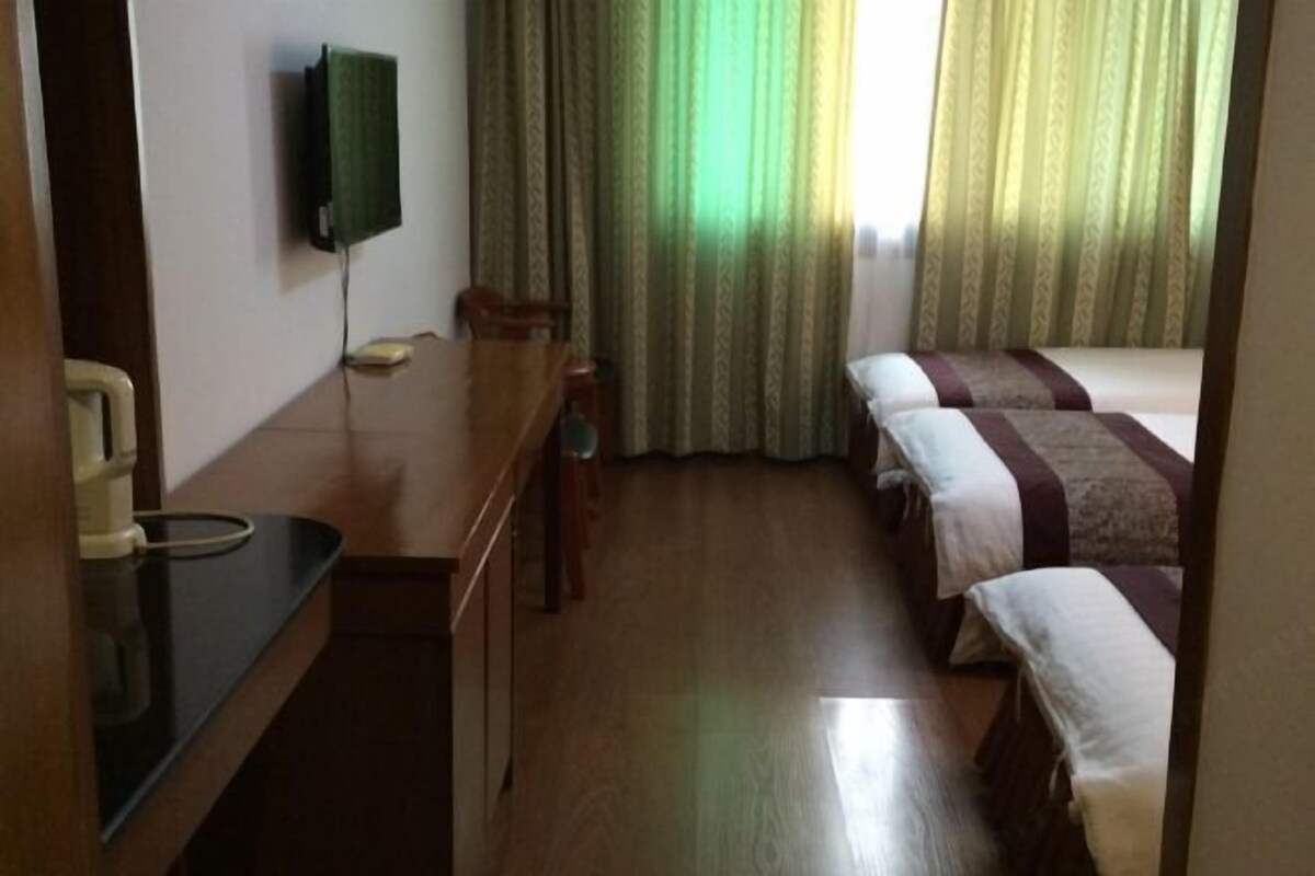 Habitación de hotel con un escritorio de madera, un televisor de pantalla plana montado en la pared, una cama con colcha a rayas y una ventana con cortinas verdes.