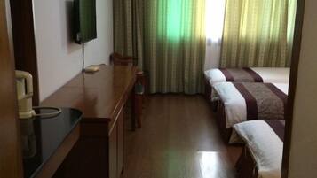 A hotel room with a wooden desk, a flat-screen TV mounted on the wall, a bed with a striped bedspread, and a window with green curtains.