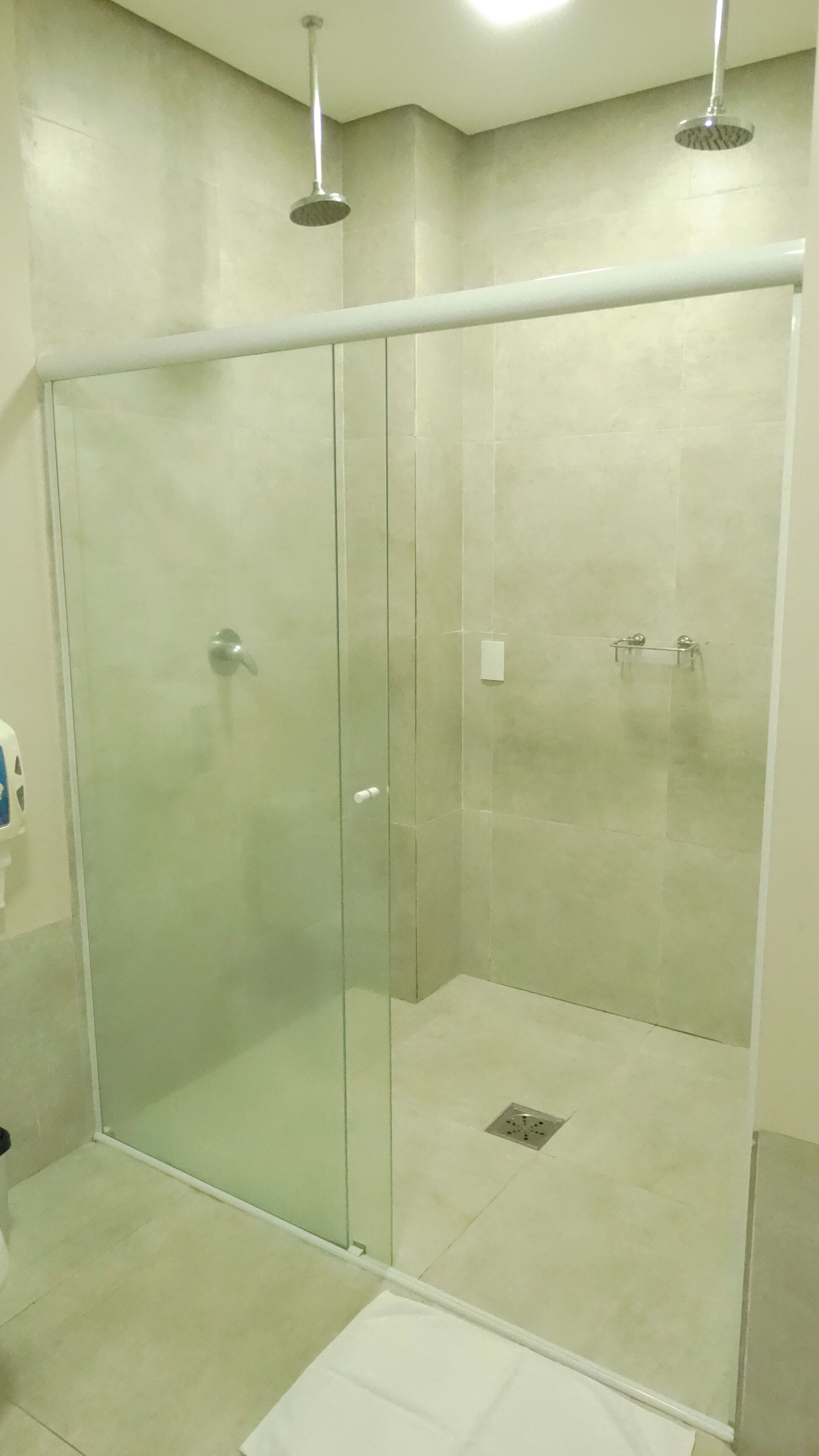 Cabina de ducha con paredes de vidrio, un tapete blanco en el suelo, un cabezal de ducha manual y un aplique en la pared.