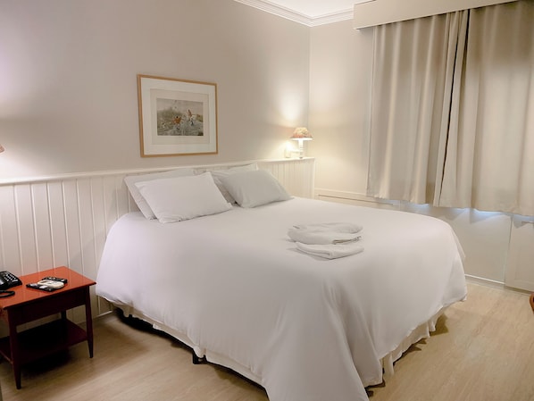 Una cama bien hecha con sábanas blancas, una mesita de noche con una lámpara y un cuadro en la pared.