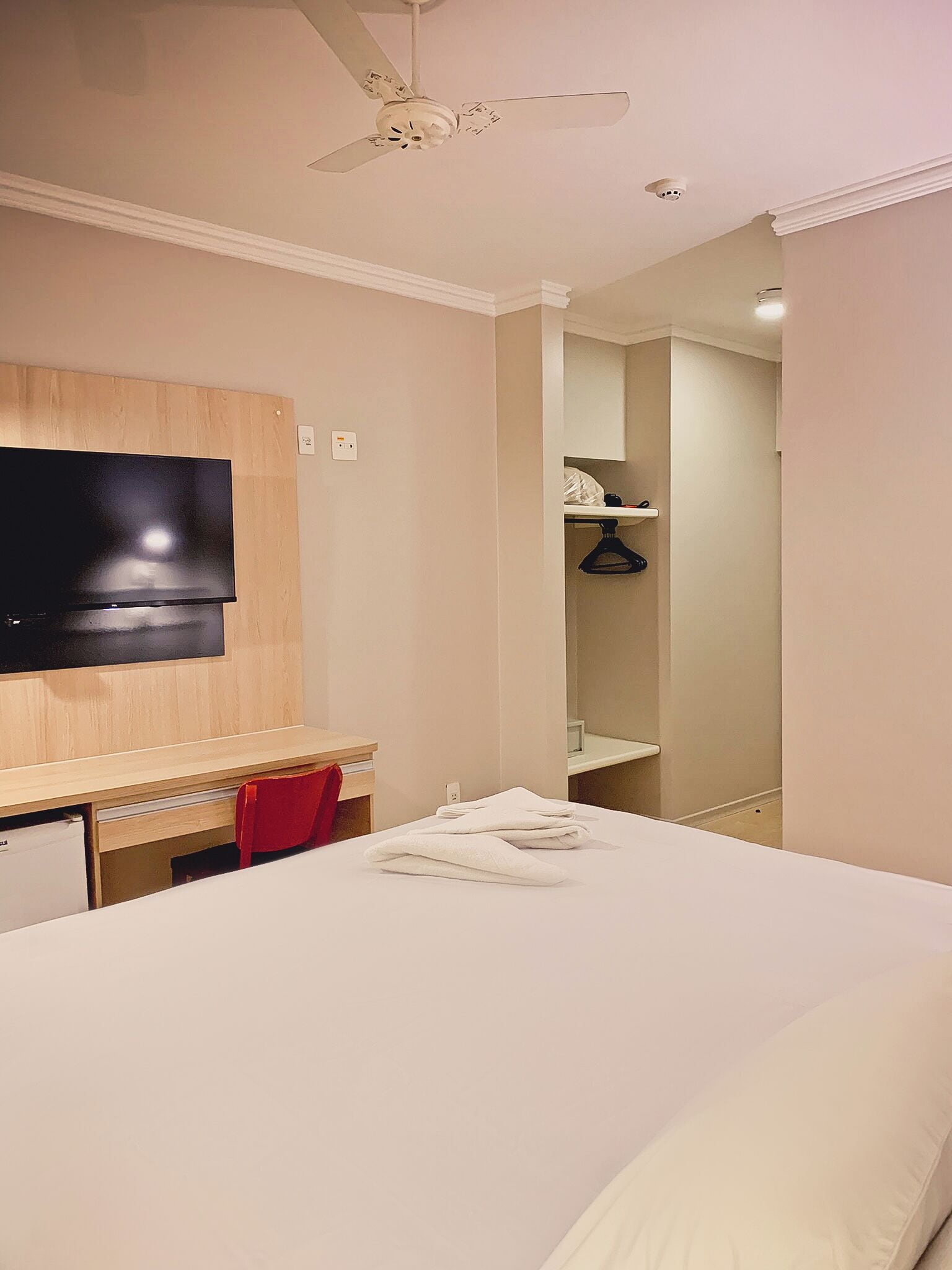 A hotel room with a bed, a desk, a TV, a ceiling fan, and a bathroom visible through an open door.