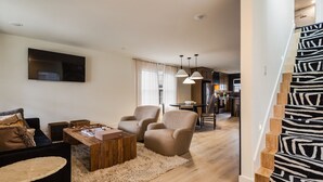A modern living room with a sofa, armchairs, a coffee table, and a TV. There is a staircase with a zebra print carpet leading to a kitchen area with a dining table and chairs. - The Winnie Lou (Jeffersonville)