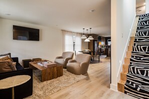 A modern living room with a sofa, armchairs, a coffee table, and a TV. There is a staircase with a zebra print carpet leading to a kitchen area with a dining table and chairs. - The Winnie Lou (Jeffersonville)