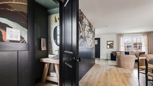 A modern interior with a dark wood door leading to a bathroom with a stone sink and a towel hanging on the wall. A living area with a sofa and chairs is visible in the background. - The Winnie Lou (Jeffersonville)