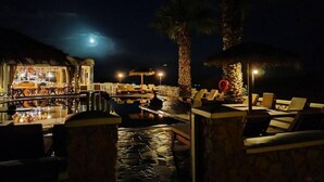 Zona de piscina nocturna con sillones de descanso y un bar.