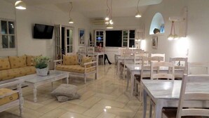 Amplia sala de estar con un conjunto de sofás, mesas de comedor y sillas. Hay un televisor montado en la pared y una planta en maceta sobre una mesa.