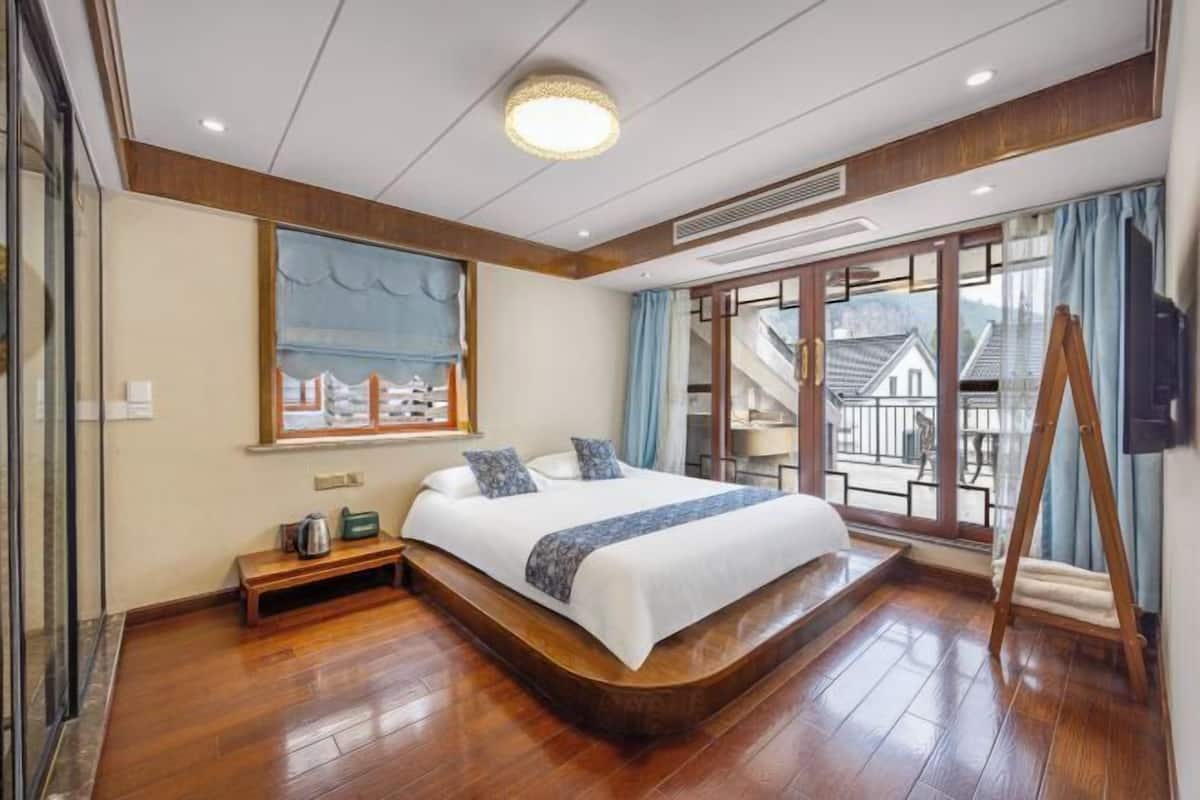 Une chambre spacieuse avec un grand lit, un plancher en bois, une vue sur une maison traditionnelle à travers une porte vitrée et une échelle en bois appuyée contre le mur.