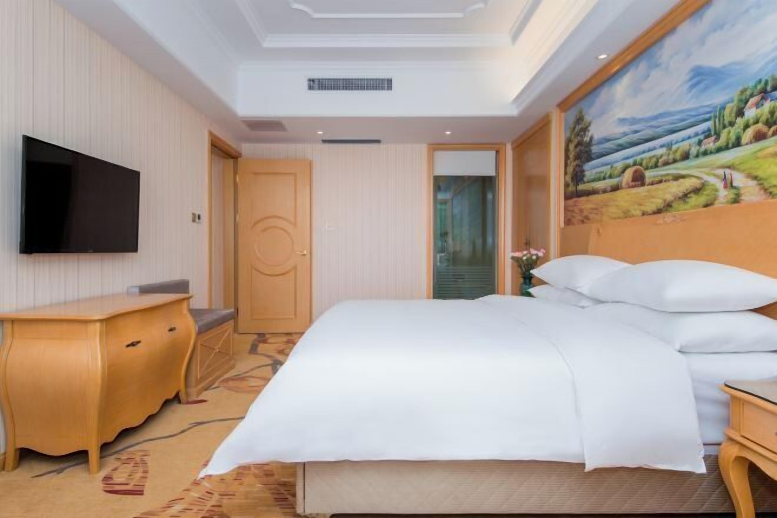 A hotel room with a bed, a TV, a wooden dresser, a painting on the wall, and a door.