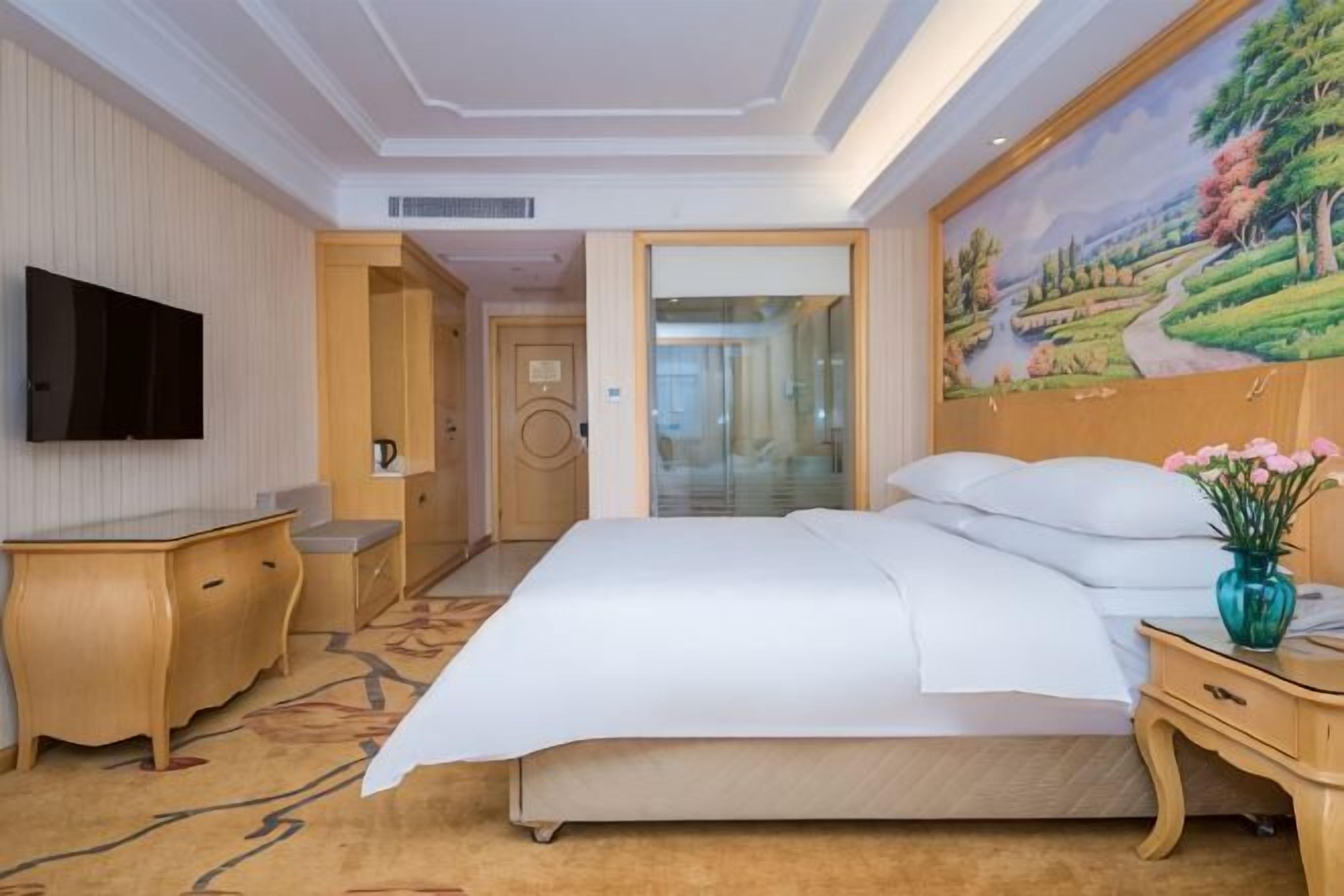 Une chambre d’hôtel avec un grand lit, un téléviseur fixé au mur, un bureau et un tableau représentant un paysage.