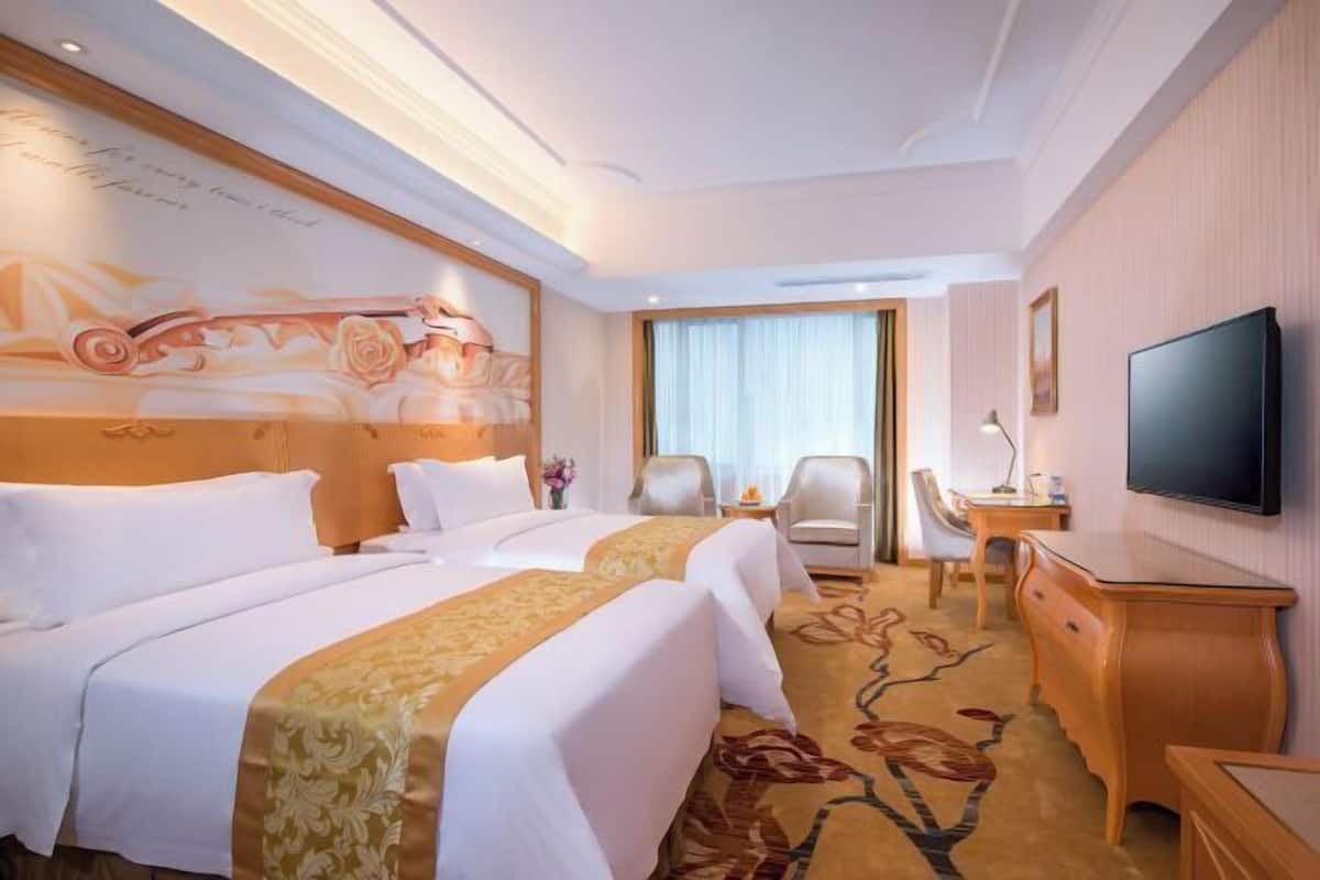 Una habitación de hotel con dos camas, un escritorio, una silla, un televisor y un adorno en la pared.