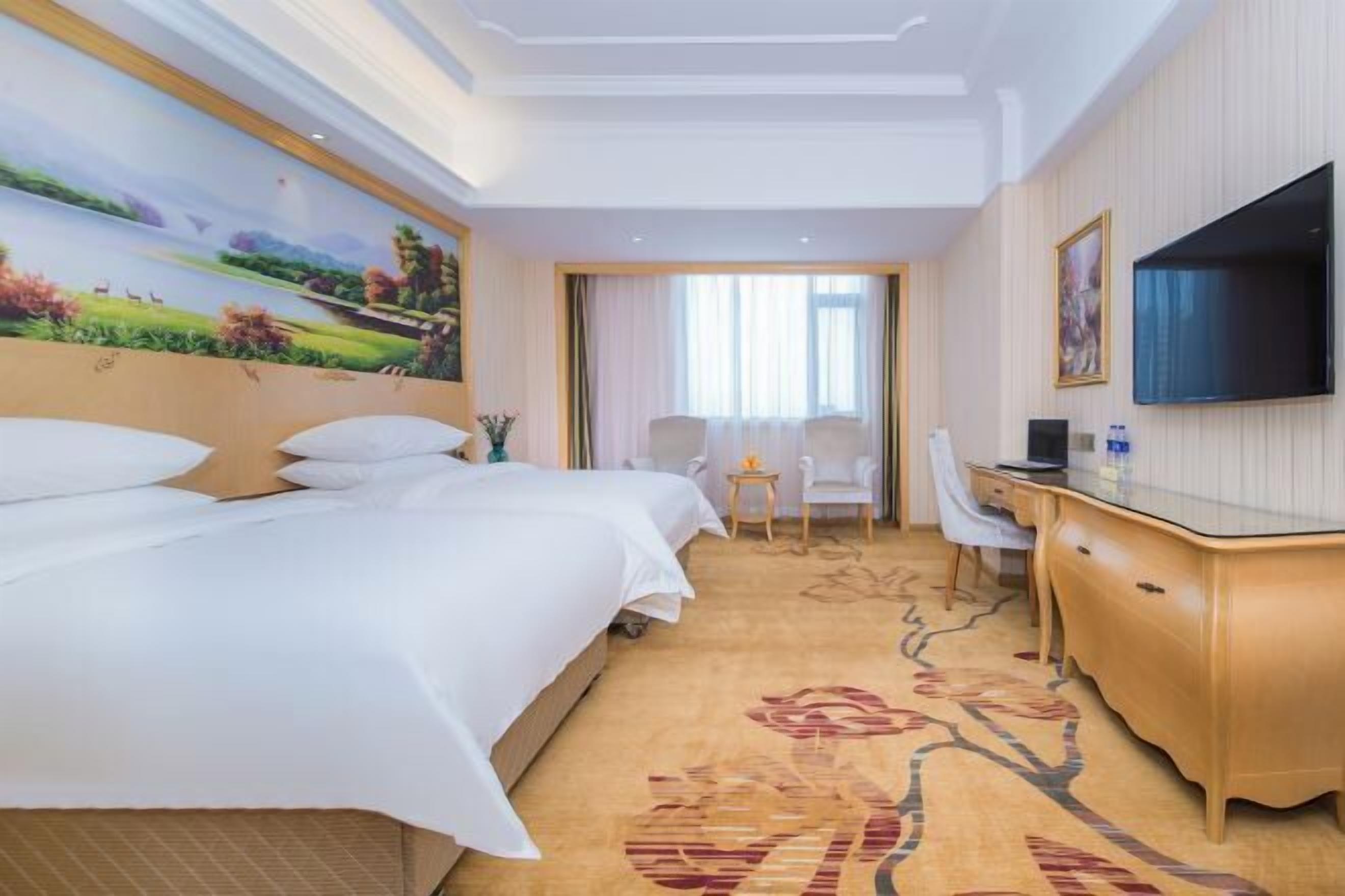 A hotel room with a large bed, a desk, a chair, a TV, and a painting on the wall.
