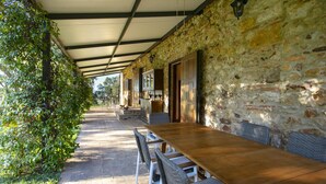 Un área de comedor al aire libre con techo, paredes de piedra, una mesa de madera larga y sillas de mimbre.