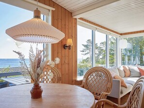 A wooden interior with a round table, wicker chairs, a sofa, and a large window overlooking the sea.