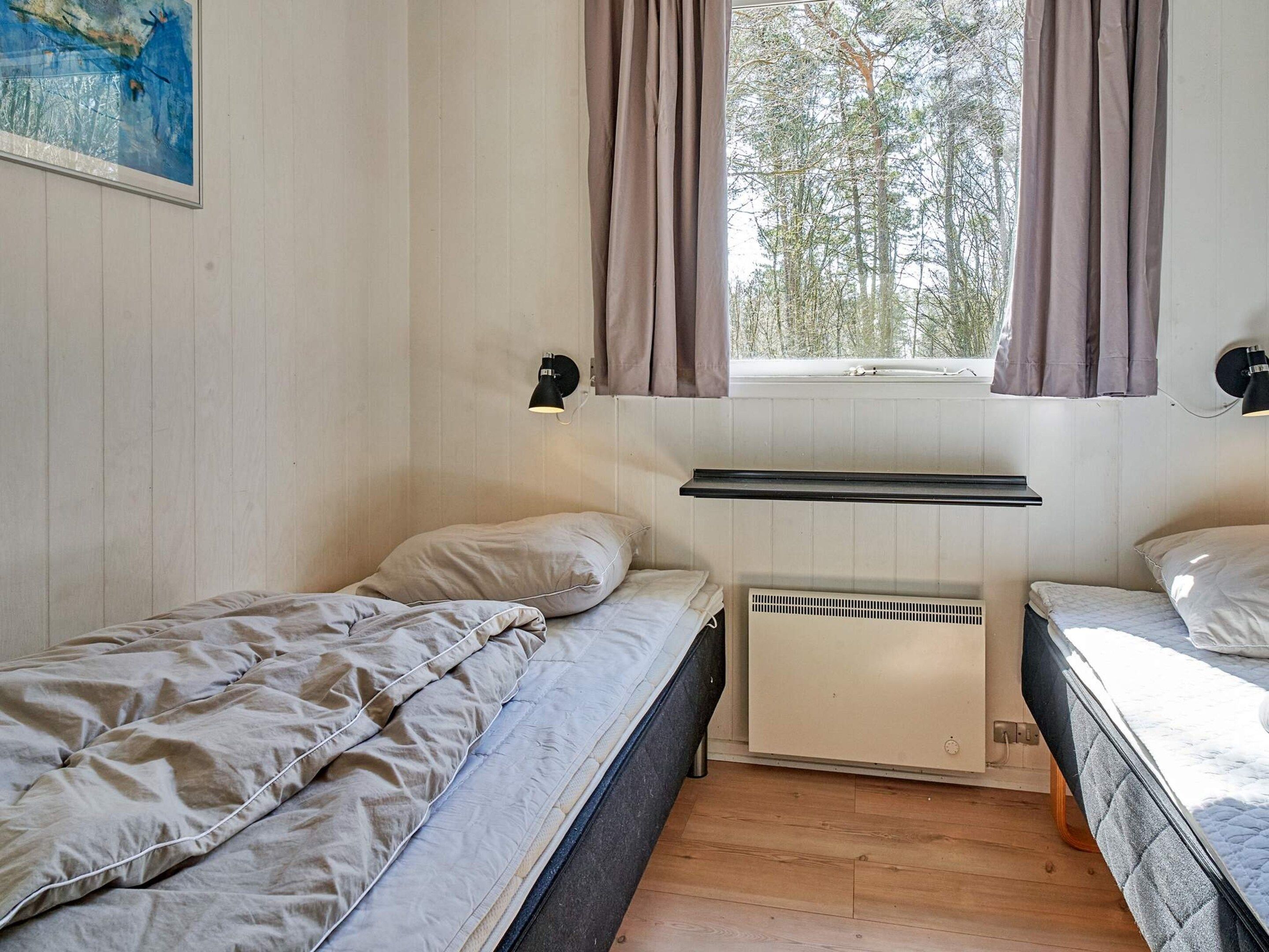 A small, single-room accommodation with two beds, a window with curtains, a radiator, and a wall-mounted light fixture.