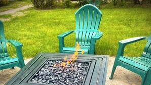 A square outdoor fire pit with a metal frame and a black surface, surrounded by green plastic chairs.