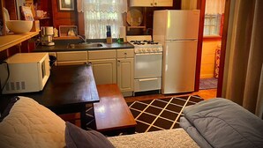 A compact kitchen with a stove, sink, and refrigerator. There is a wooden table and a couch in the foreground.