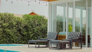 Un área junto a la piscina con sillones de descanso, una pérgola de madera y un ventanal que da a un espacio interior.