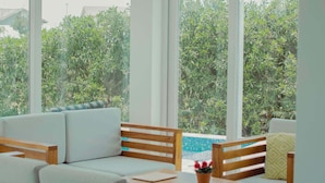 Un salón moderno con sofá, sillas de madera, una mesita y vistas a una piscina y zonas verdes.