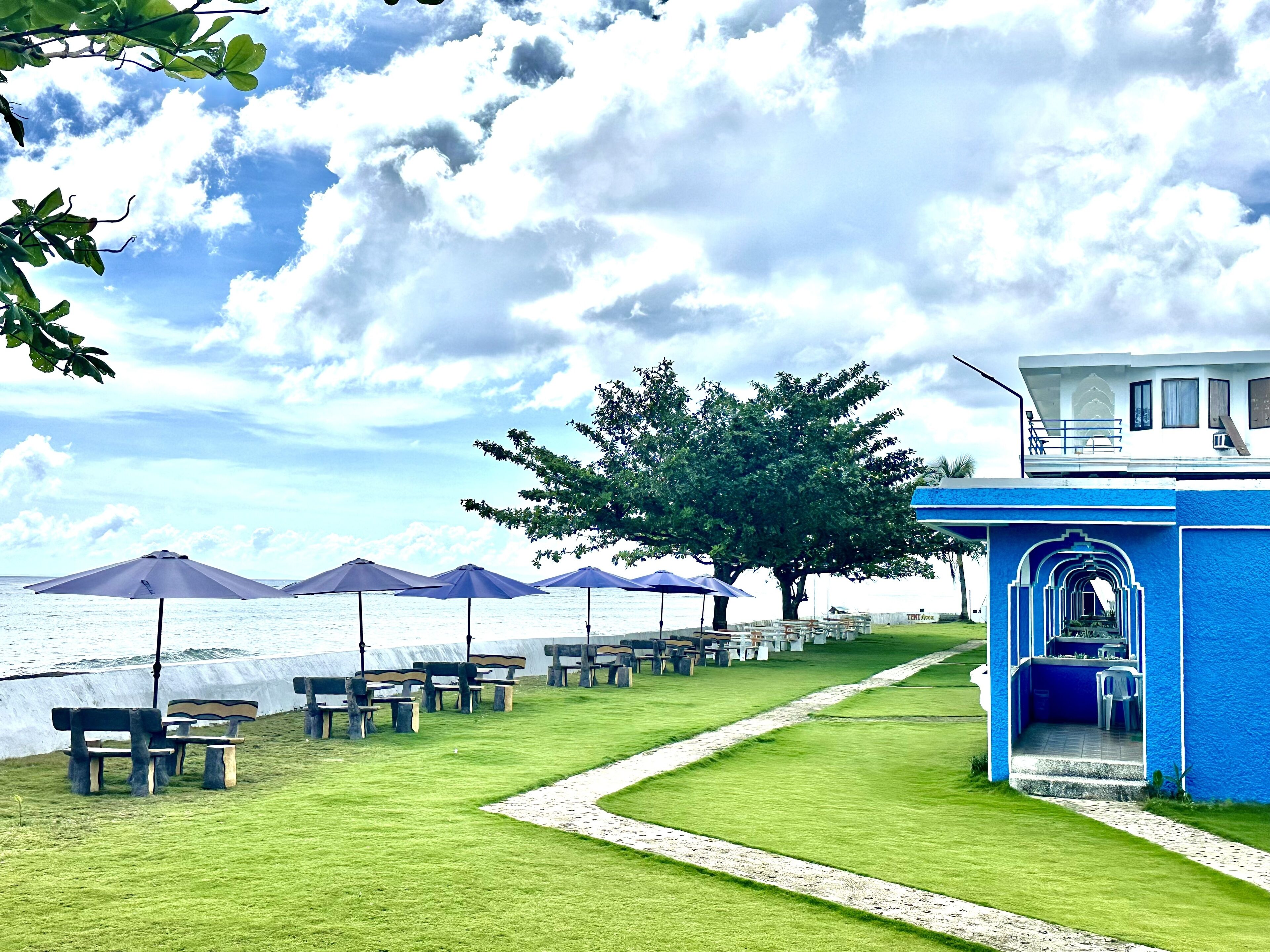 A beachside area with a pathway leading to a blue building with an archway entrance, surrounded by benches and umbrellas.