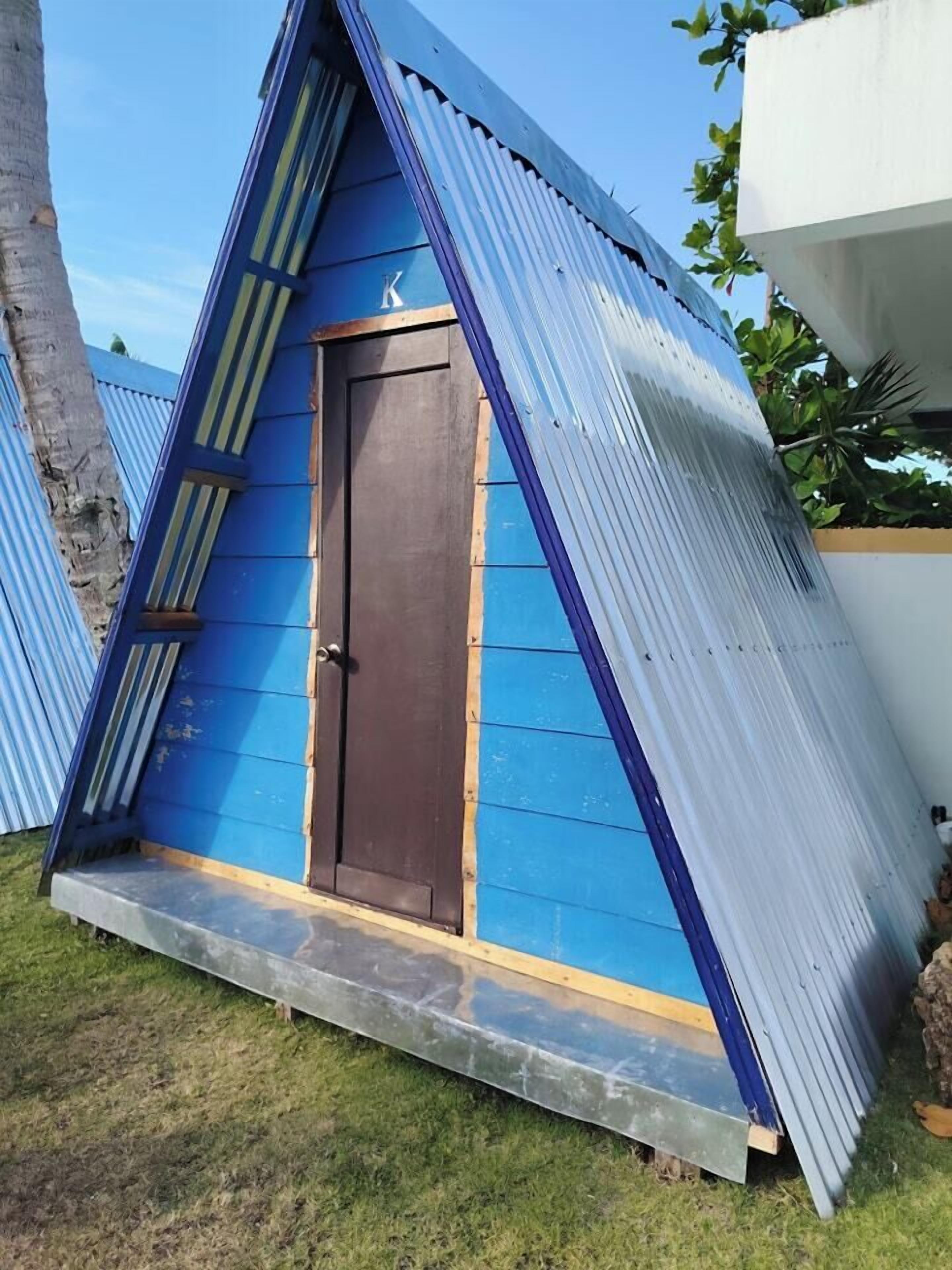 A small, blue, triangular structure with a dark brown door, surrounded by grass and metal sheets.