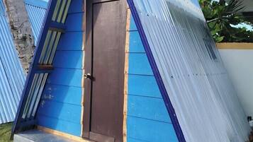 A small, blue, triangular structure with a dark brown door, surrounded by grass and metal sheets.