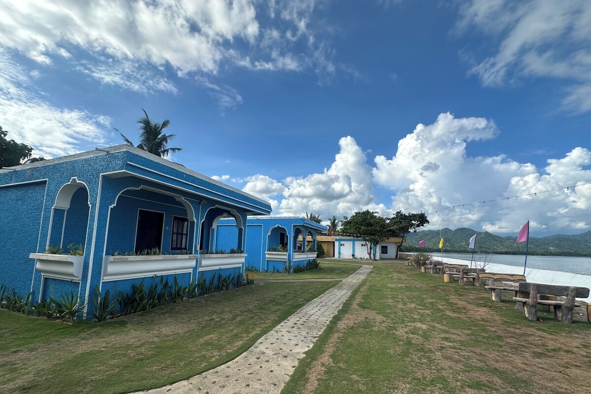 A blue building with arched windows and a pathway leading to a seating area by a body of water.