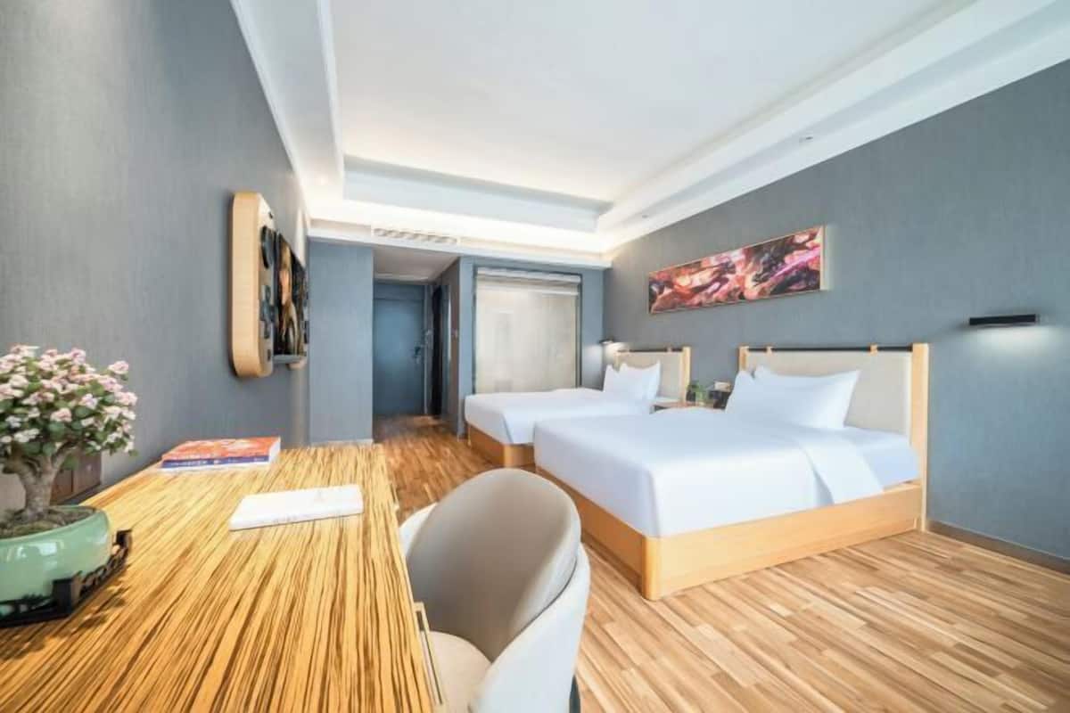 Une chambre d’hôtel avec deux lits, un plancher en bois, un bureau avec une chaise, une petite plante et un tableau au mur.
