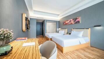 A hotel room with two beds, a wooden floor, a desk with a chair, a small plant, and a painting on the wall.