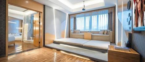 A modern hotel room with a wooden floor, a sofa bed, a desk, and a large window with curtains.