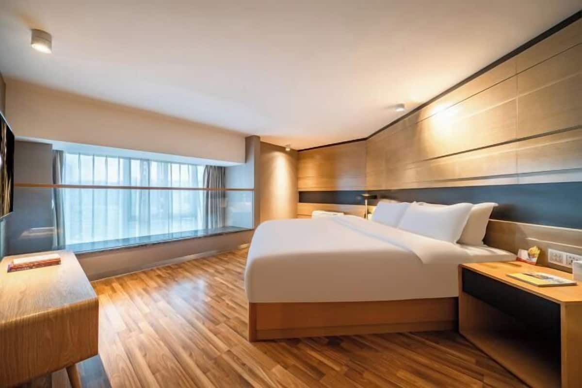 Une chambre d’hôtel moderne avec un grand lit, un plancher en bois, une table de chevet, une lampe de chevet et une grande fenêtre avec des rideaux.