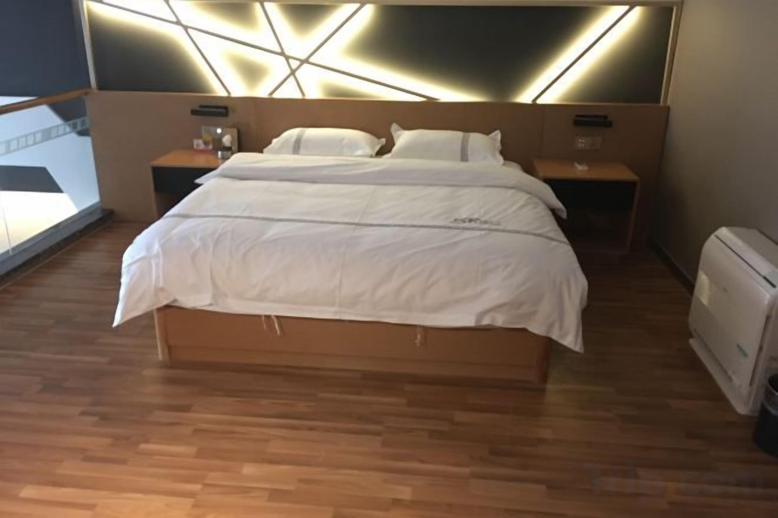 A modern hotel room with a large bed, wooden flooring, a headboard with integrated lighting, a nightstand, and a bedside table.