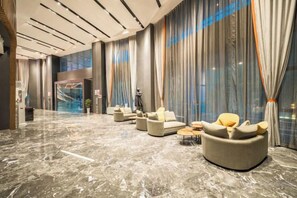 Un lobby moderno con piso de mármol, zona de estar con sofás y sillones, ventanas amplias con cortinas y una escalera al fondo.