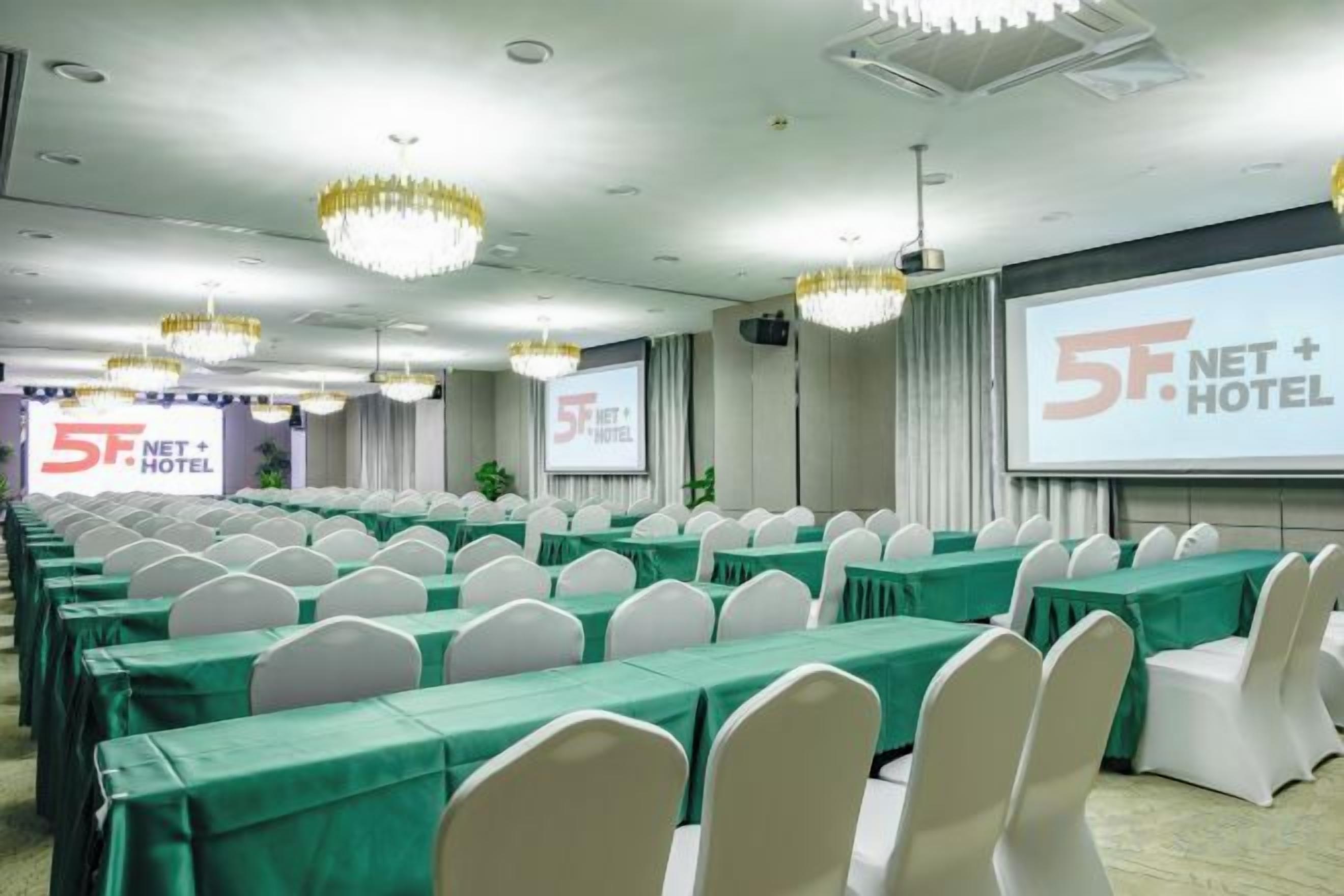 Uma sala de conferências com fileiras de mesas e cadeiras, uma tela grande e lustres.