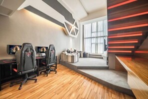 Una sala de juegos moderna con dos sillas gamer, una pantalla grande y una zona de estar acogedora con una escalera que lleva a un balcón.