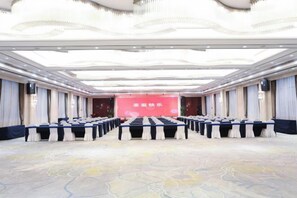 Une salle de conférence avec des rangées de tables et de chaises, une scène dotée d’un fond rouge et un tapis à motifs.