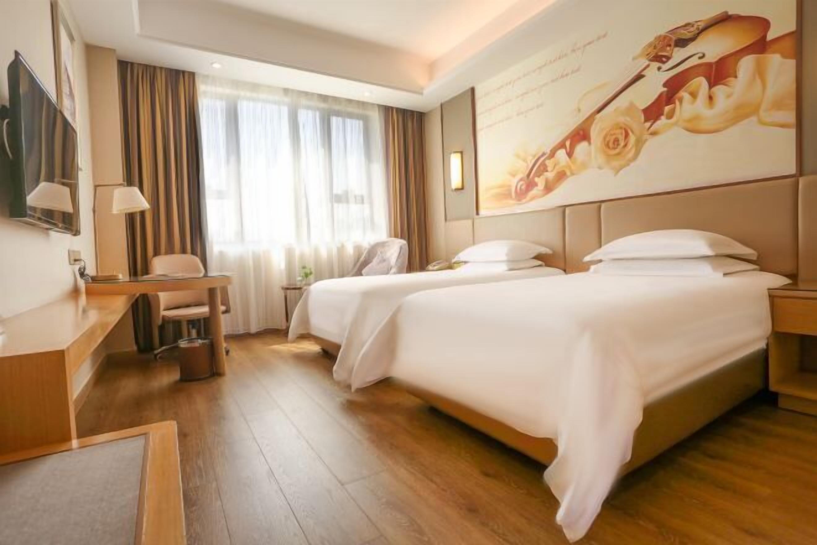 A hotel room with two beds, a desk, a chair, a TV, and a large window with curtains.