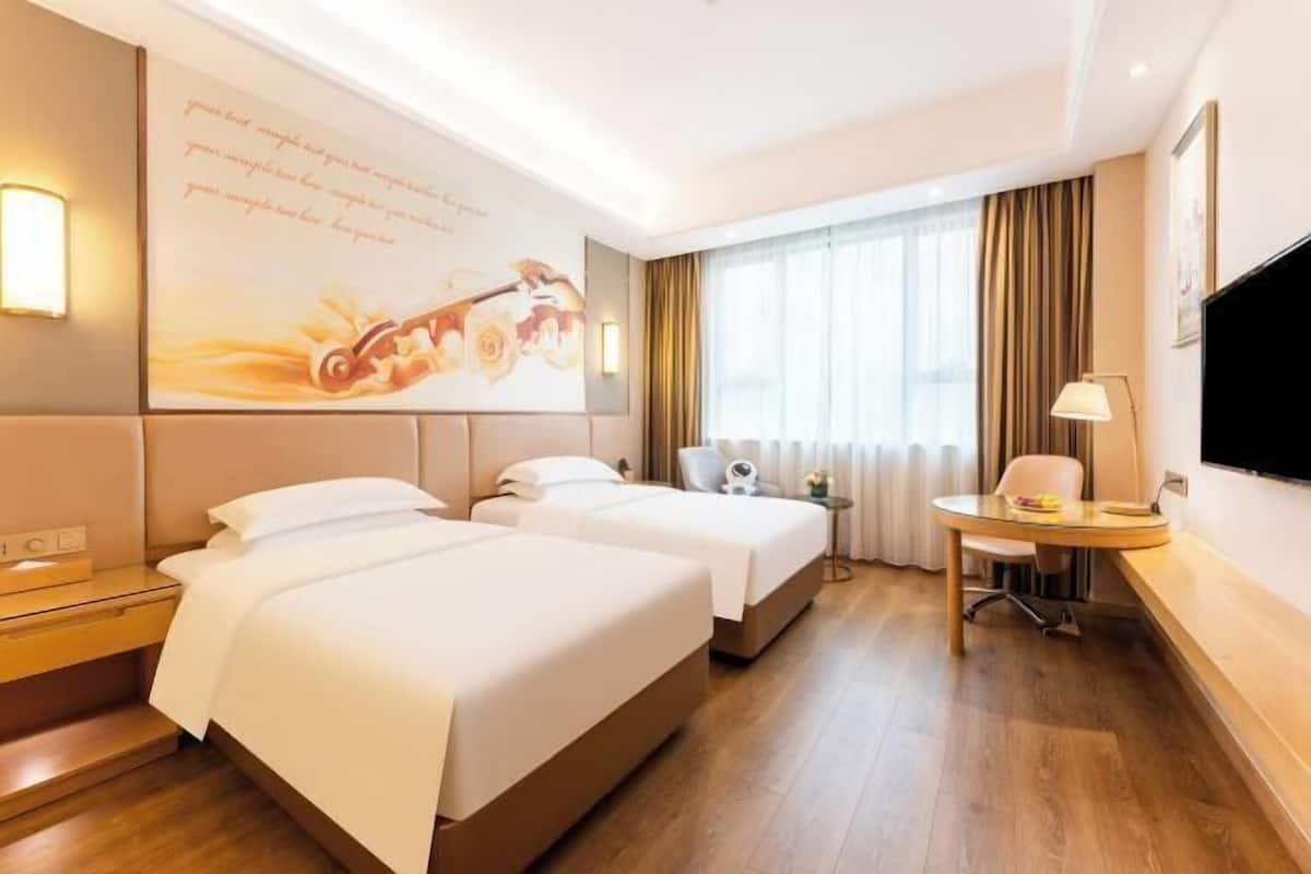 Habitación de hotel con dos camas, un escritorio, una silla, un televisor y un ventanal con cortinas.