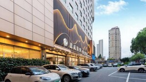 A hotel with a modern facade, a parking lot with several cars, and a building with a wave-like design on the side. - Vienna International Hotel (Wenzhou Avenue Railway Store) (Wenzhou)