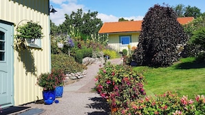 A yellow building with a blue door, a garden with various plants and flowers, and a red-tiled roof house in the background. - 2 Person Holiday Home in Brastad-by Traum (Brastad)