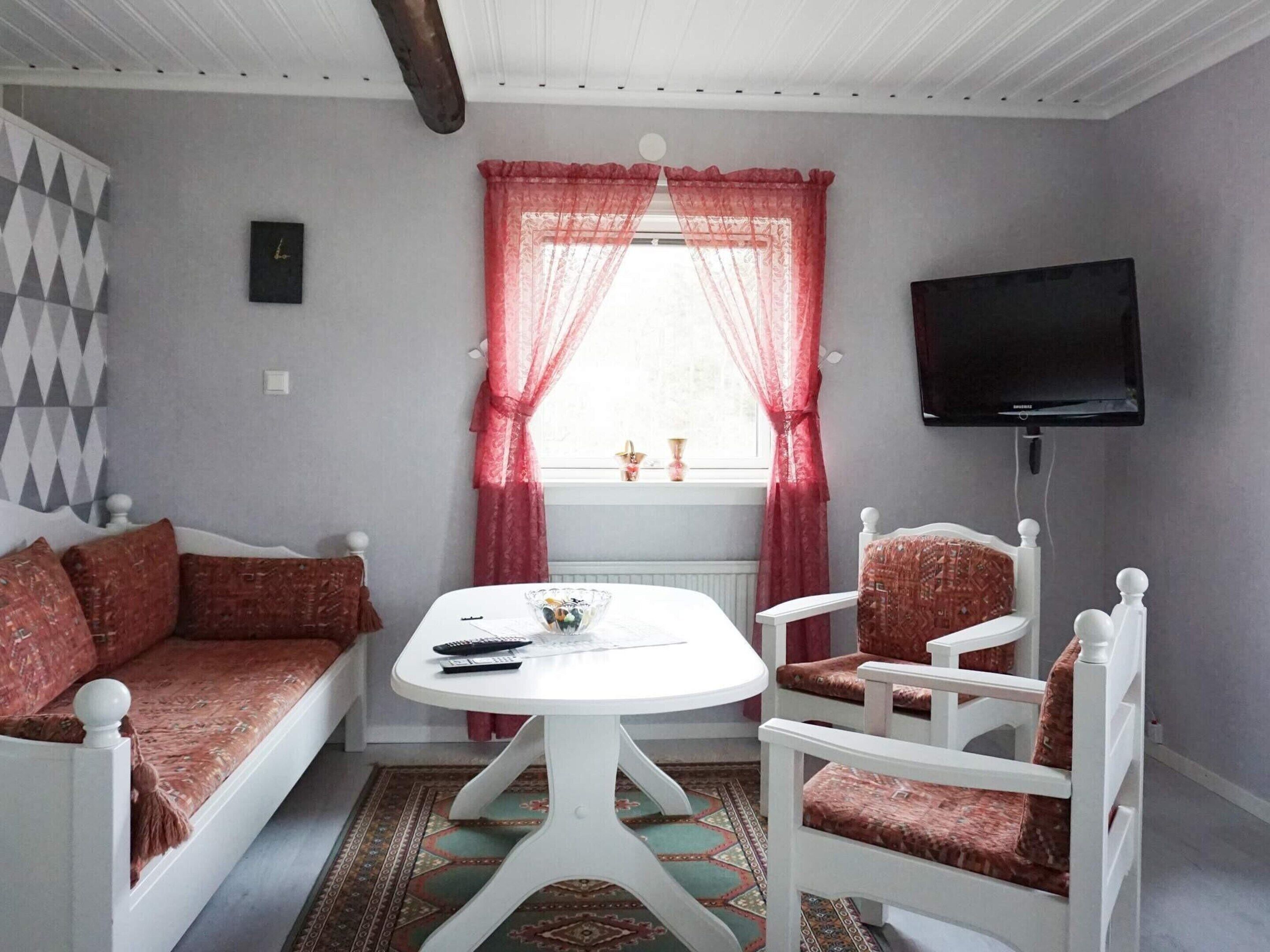 Una habitación con un sofá blanco, dos sillas blancas, una mesita redonda con un cuenco y mandos a distancia, un televisor en la pared y una ventana con cortinas rojas.