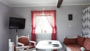 A cozy room with a white sofa, two white chairs, a small round table, a TV mounted on the wall, and a window with red curtains. - 2 Person Holiday Home in Brastad-by Traum (Brastad)