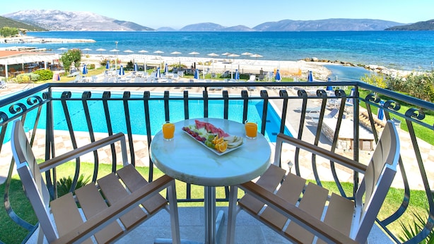 A balcony with a pool, a table with a fruit platter and drinks, and a view of the sea and mountains. - Sami Beach Hotel (Kefalonia)
