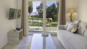 A modern living room with a sofa, a coffee table, a TV mounted on the wall, and a view of a patio with outdoor furniture. - Sami Beach Hotel (Kefalonia)