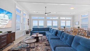 A spacious living room with a large blue sectional sofa, a coffee table, and a dining area with wooden chairs and tables. - Money Beach: Private Pool, 7 Bedrooms, Sleeps 25! (Indian Pass)