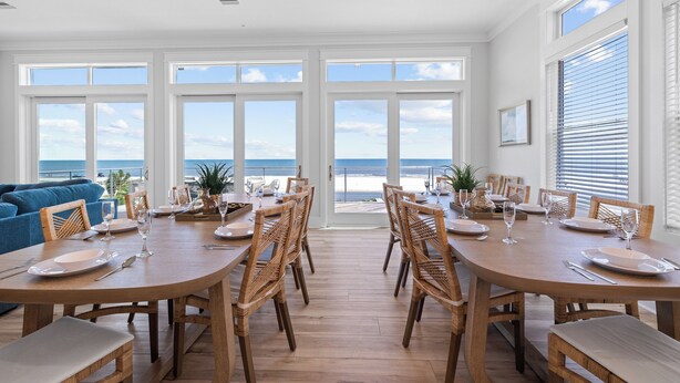 A dining area with a view of the ocean, featuring a wooden table set for six, wicker chairs, and large windows. - Money Beach: Private Pool, 7 Bedrooms, Sleeps 25! (Indian Pass)