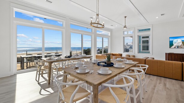 A spacious dining area with a wooden table and white chairs, large windows offering a view of the beach, and a modern chandelier.
