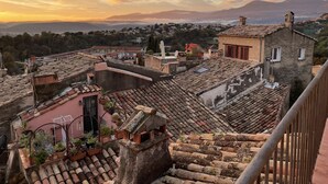 A rooftop terrace with terracotta tiles, potted plants, and a view of a town at sunset.